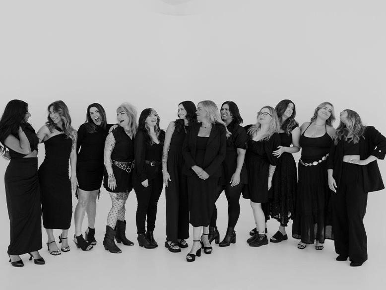 A group of twelve women stand together in a studio, all dressed in black outfits, posing and smiling for the camera against a plain white background.