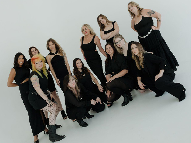 A group of thirteen women dressed in black pose together against a plain white background.