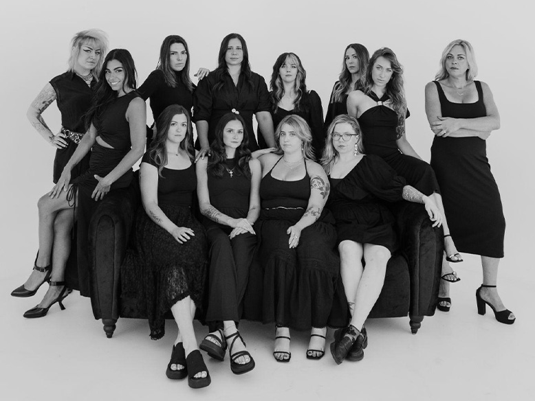 A group of thirteen women pose together in black outfits, with some seated on a couch and others standing behind, against a plain white background.